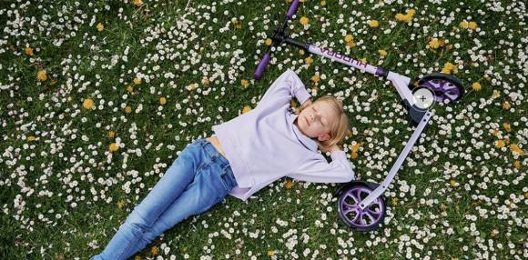 Kind liegt in Blumenwiese mit Scooter neben sich 