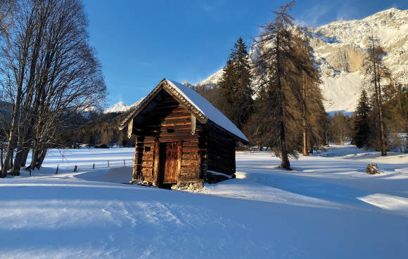 Winterlandschaft mit Bergen im Hintergrund und einer Holzhütte und Bäumen im Vordergrund 